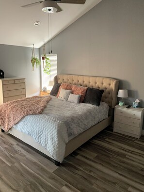 Ein Schlafzimmer mit einem gesteppten Kopfteil, einem Bett mit weißer Steppdecke, einer flauschigen rosafarbenen Decke, einem Nachttisch mit Lampe und einer hängenden Pflanze.