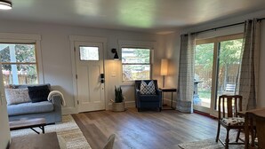 Un vaste espace de vie comprenant un canapé gris, une table basse en bois, un fauteuil et une table à manger. Il y a deux fenêtres avec des rideaux, une lampe et une plante en pot.