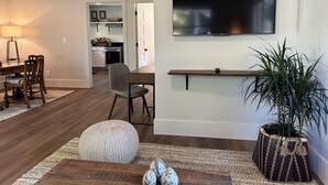 Un salon avec un téléviseur fixé au mur, un coin repas avec des chaises et une table, et une cuisine à l’arrière-plan.