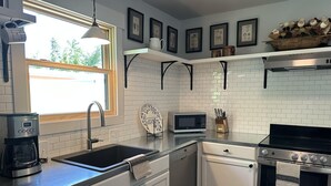 Une cuisine moderne avec des meubles blancs, un évier en acier inoxydable, un four à micro-ondes, un lave-vaisselle et une cuisinière. Des tableaux encadrés ornent le mur, et une fenêtre offre une vue sur l’extérieur.