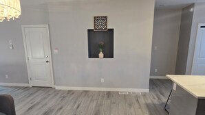 Una habitación amplia con paredes grises, piso de madera, una puerta blanca, un tapete negro y un estante pequeño con un florero.
