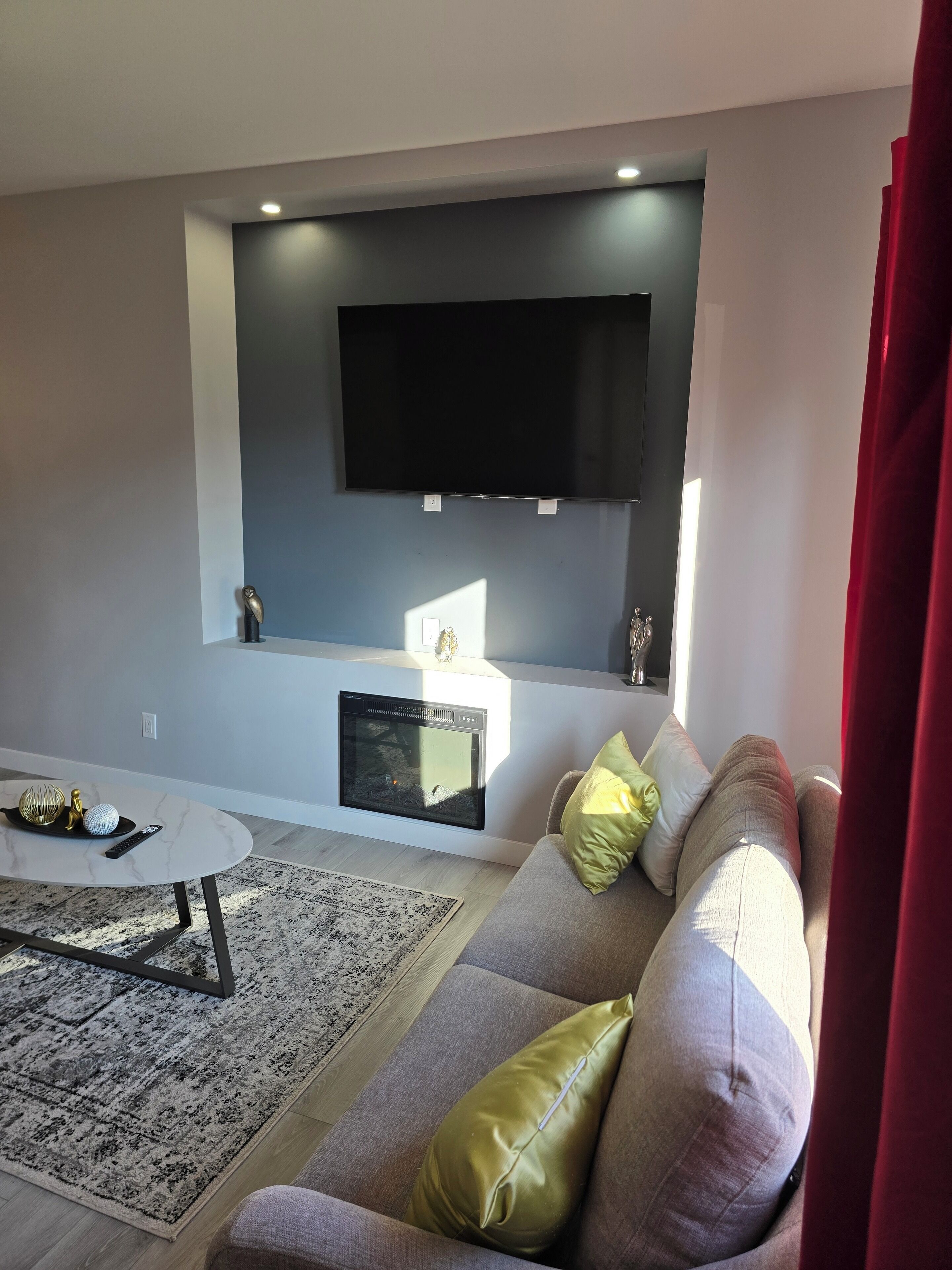 Een moderne woonkamer met een flatscreen televisie aan de muur, een open haard onder de televisie, een bank met kussens, een salontafel en een kamerbreed tapijt met een patroon.