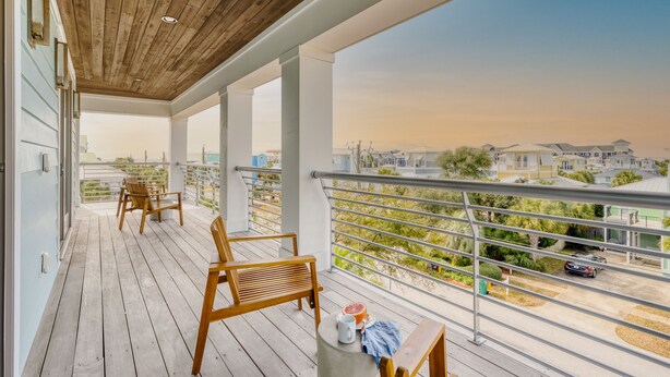 Un terrazzo in legno con sedie e un tavolo, affacciato su una zona residenziale con case e cielo sereno.
