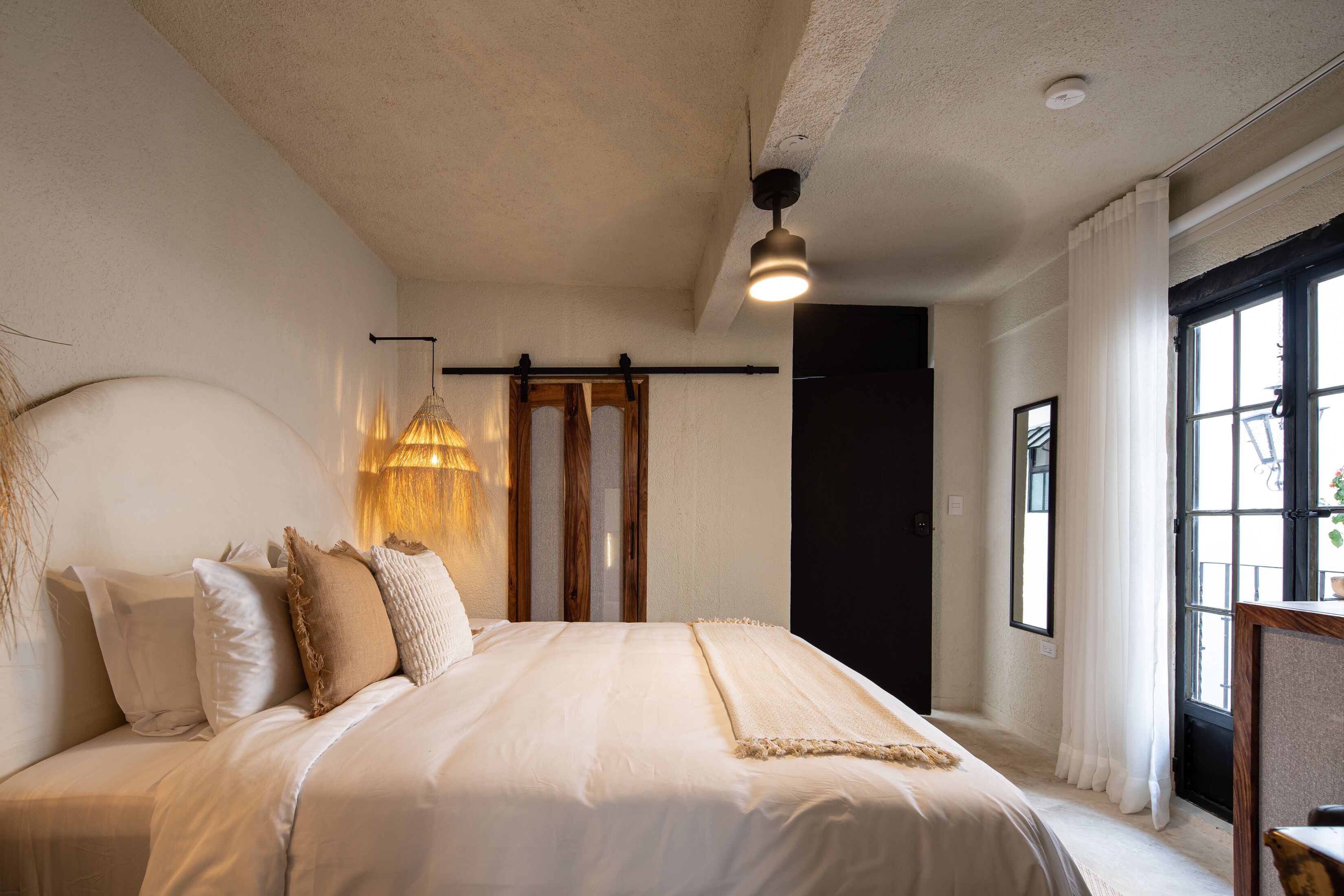Un dormitorio con una cama grande, un lámpara colgante, una ventana con cortinas y una puerta.