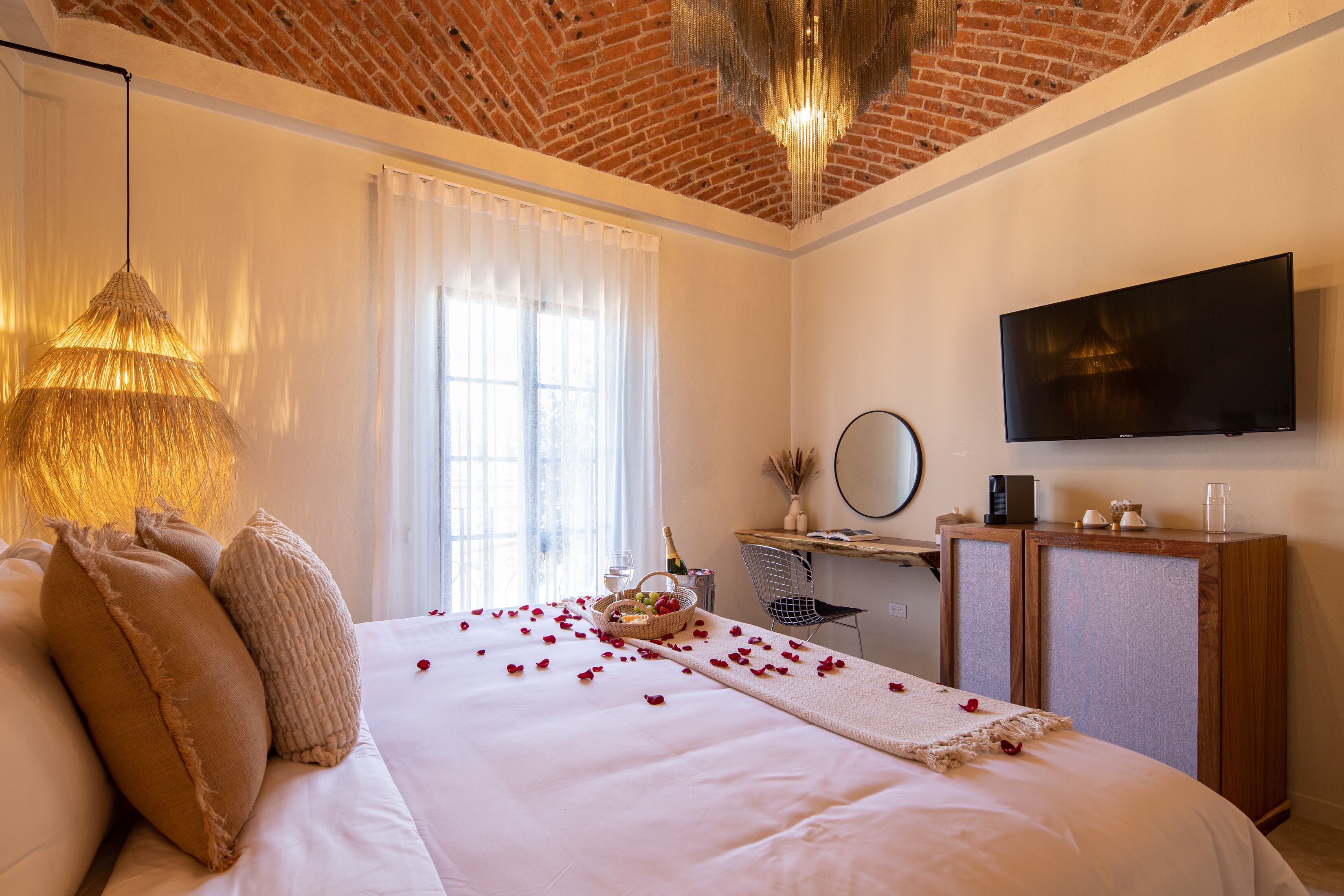Une chambre d’hôtel avec un plafond en briques, un grand lit parsemé de pétales de roses, un lustre, une télévision et un bureau sur lequel se trouve une bouteille de champagne.