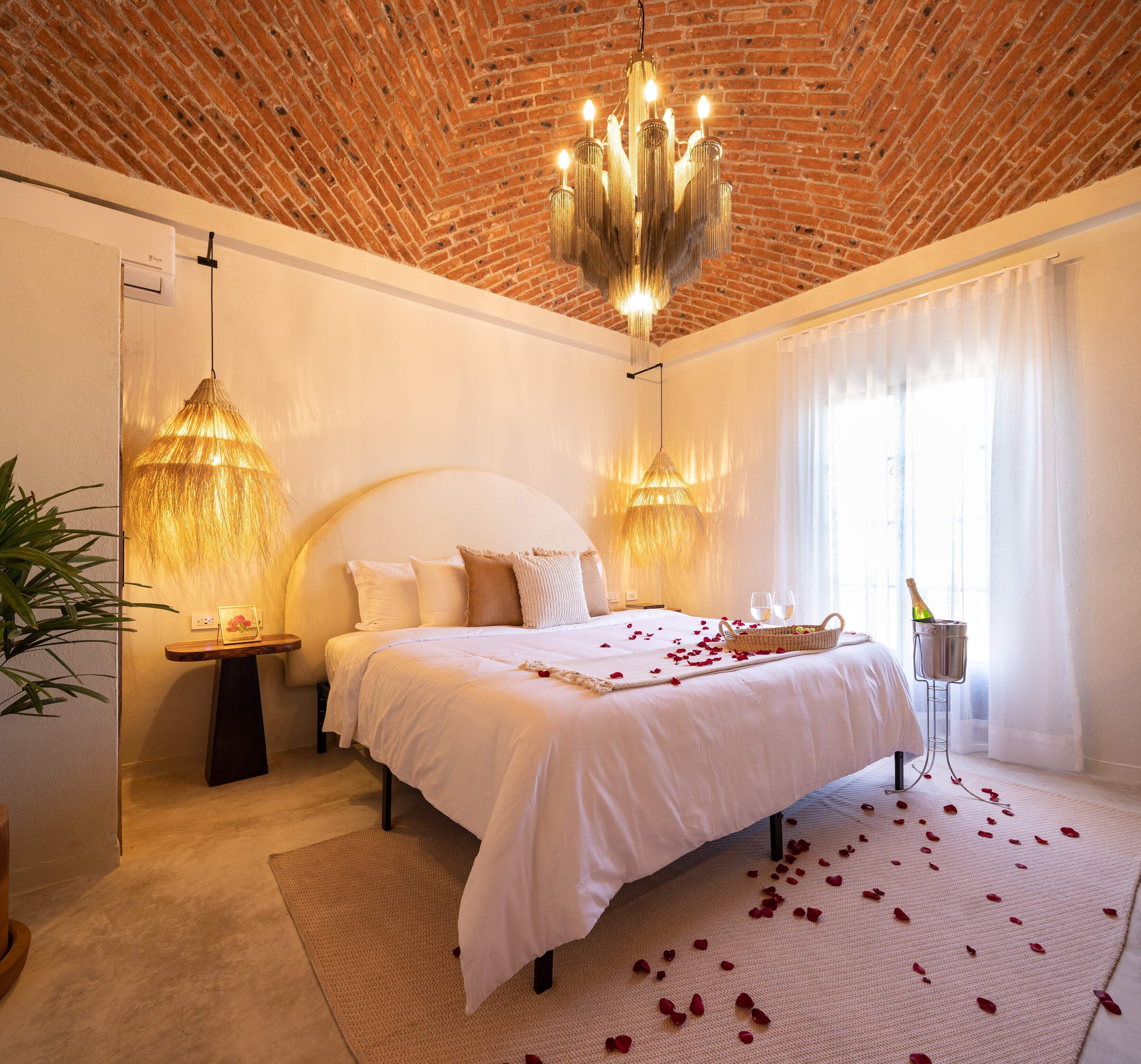 Habitación de hotel con techo de ladrillo, una cama grande con ropa blanca, dos lámparas colgantes y una botella de champán sobre una mesita de noche.