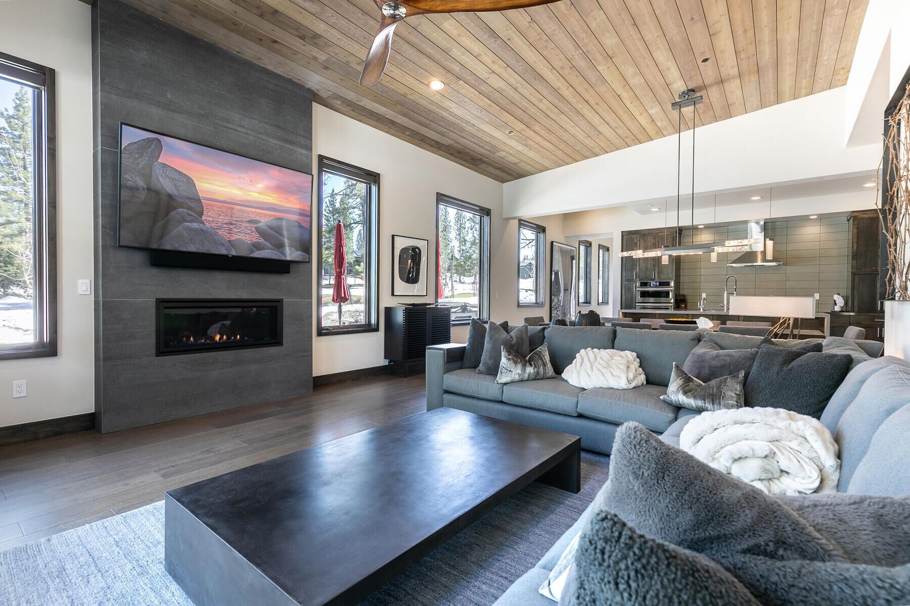 A modern living room with a fireplace, large windows, a sofa, and a kitchen area.