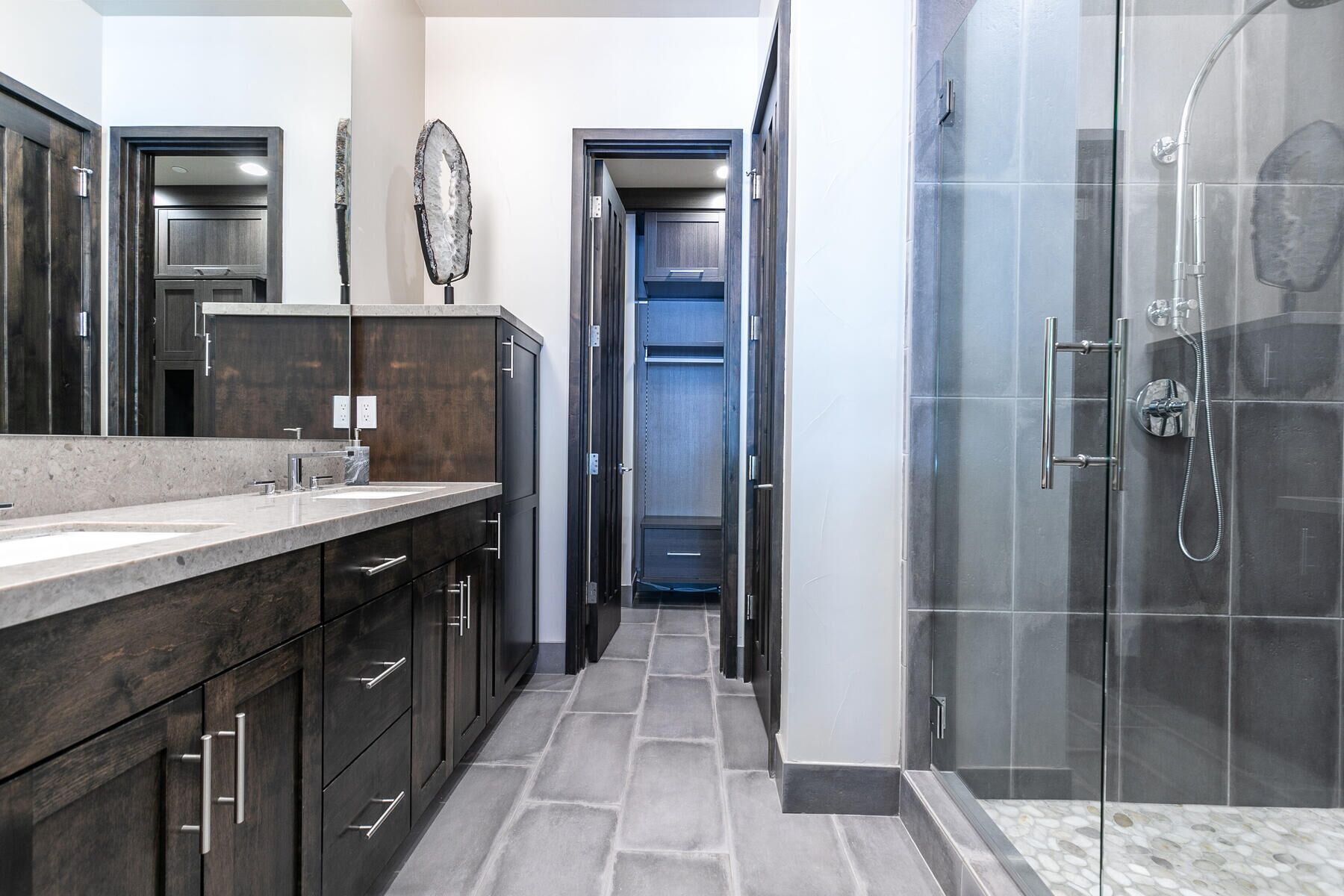 A modern bathroom with a large mirror, dark wooden cabinets, a shower with a glass door, and a tiled floor.