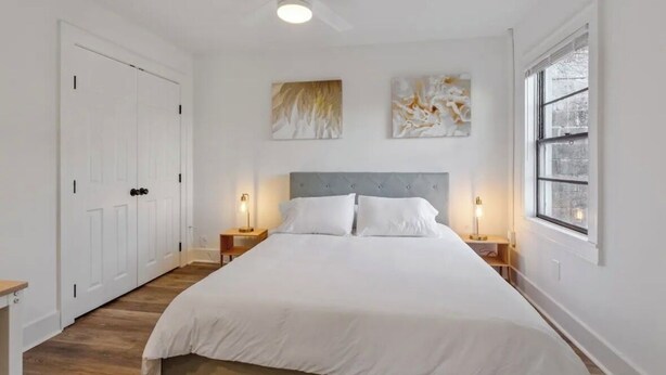 Un dormitorio con una cama grande, dos mesitas de noche con lámparas, un ventilador de techo y un cuadro abstracto en la pared.
