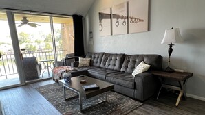 Ein modernes Wohnzimmer mit einer Eckcouch, einem Couchtisch, einem Schreibtisch und einer Lampe. Ein großes Fenster bietet einen Blick ins Freie.