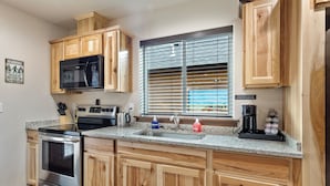 Una cocina con armarios de madera, estufa de acero inoxidable, microondas, lavaplatos y una ventana con persianas.