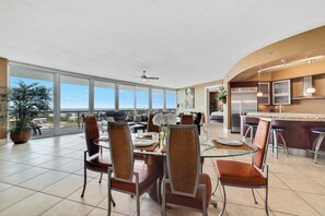 A spacious dining area with a glass table and chairs, a ceiling fan, and a view of the city. - Bella Luna 1006 - Dream Vacation-Upscale Large Luxury Condo-Sleeps 10 (Orange Beach)