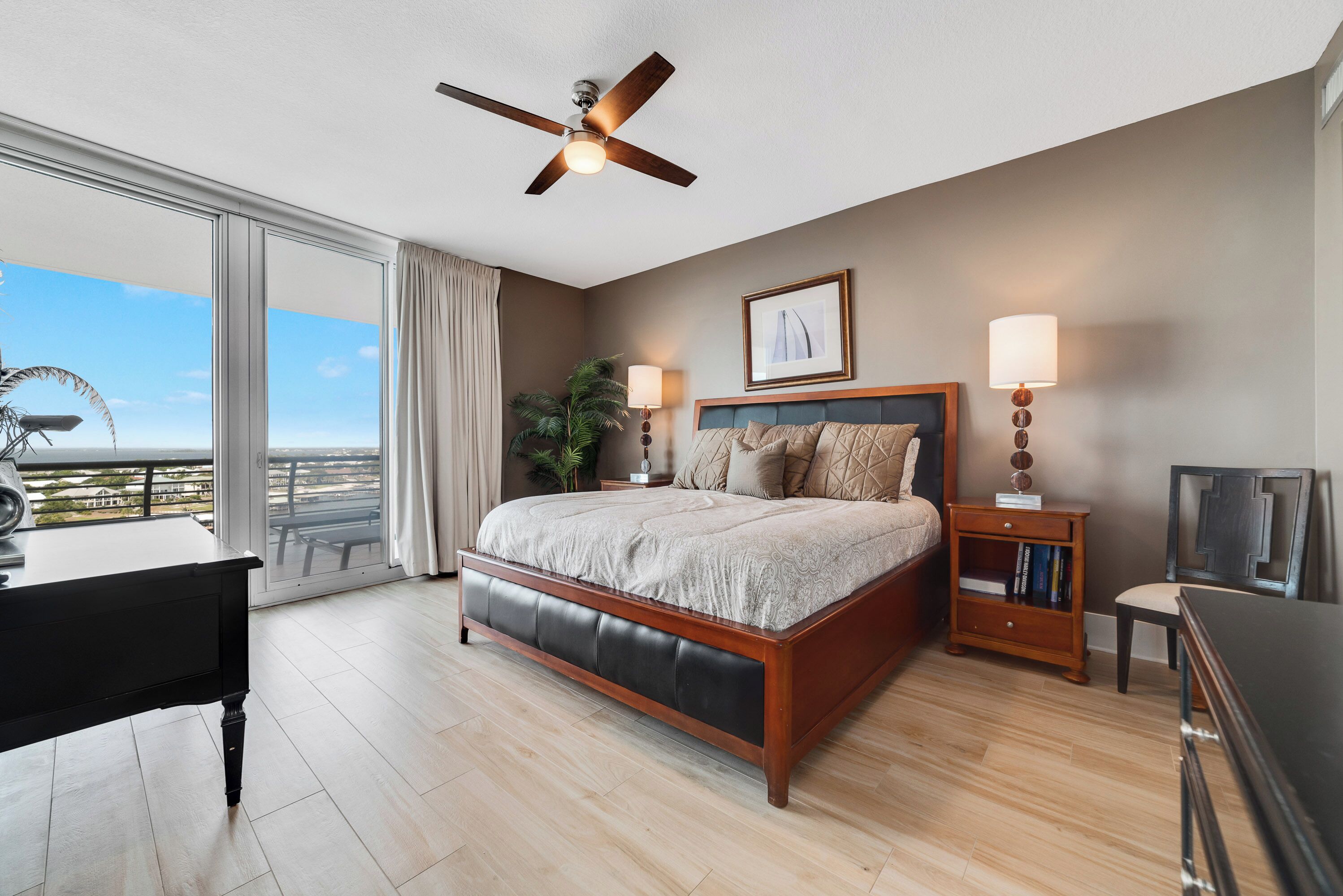 A bedroom with a bed, a desk, a ceiling fan, a nightstand, a lamp, and a view of the outdoors.