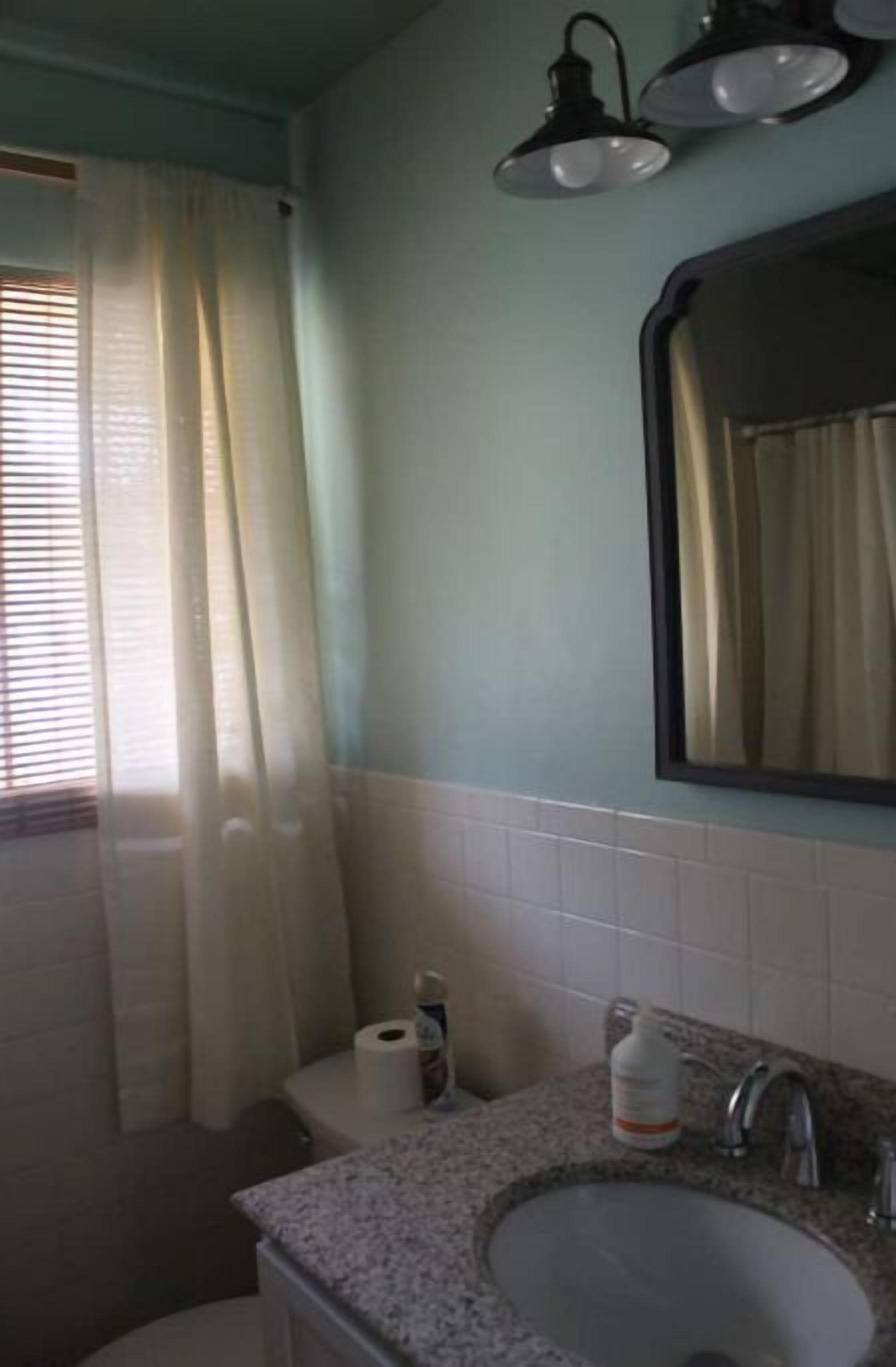 Een badkamer met een wastafel, toilet en spiegel. Boven de spiegel hangen twee lampen en er is een raam met gordijnen.