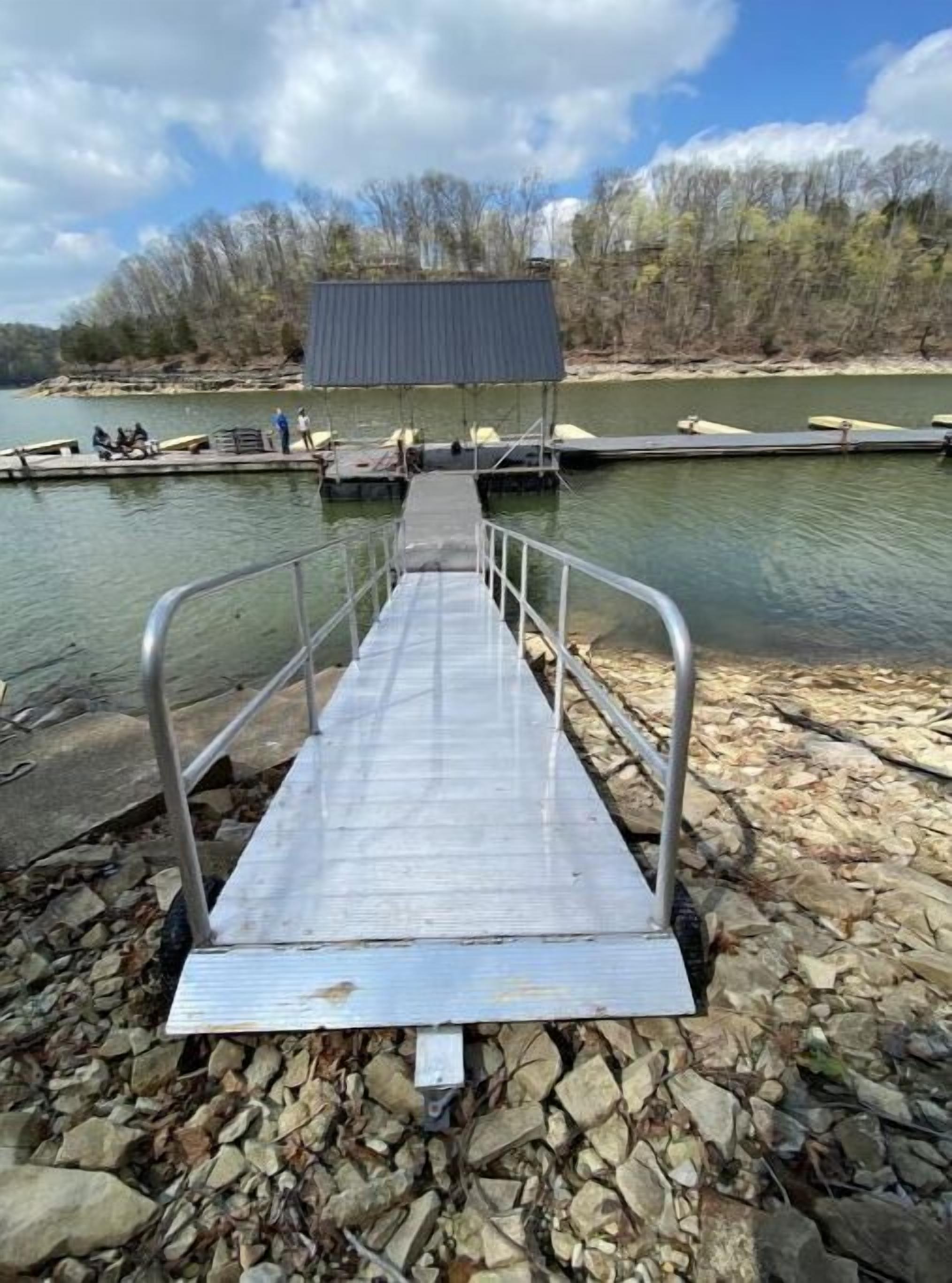 Une jetée métallique menant à une zone abritée au bord de l’eau, entourée de rochers et d’arbres.