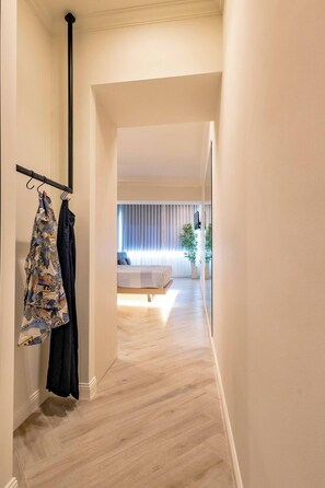 Un couloir mène à une chambre à coucher qui contient un lit, une fenêtre avec des stores et un porte-manteau mural sur lequel sont suspendus deux vêtements.