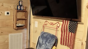 Interior de madeira com televisão fixada, uma prateleira com enlatados, uma bandeira e um banco.
