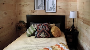 Quarto com painéis de madeira, uma cama, almofadas, uma luminária e duas obras de arte emolduradas na parede.