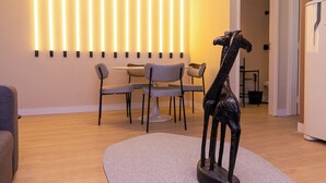 Un salon moderne avec un coin repas, agrémenté d’une sculpture de girafe originale posée sur un tapis, d’un luminaire fixé au mur et d’un réfrigérateur blanc.