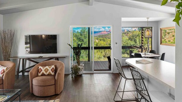A modern living room with a TV, comfortable seating, a dining area with a bar, and large windows offering a view of the outdoors.