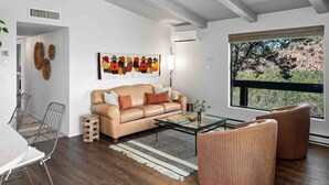 A modern living room with a beige sofa, a glass coffee table, and a large window offering a view of trees and hills.