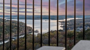A view through a window with vertical bars, overlooking a lake, surrounding forest, and distant buildings. - Timeless Lake Views, Indoor Pool, Game Room, Hot tub, Firepit, Minutes from SDC (Branson)