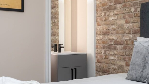 A bedroom with a brick wall, a white bed, a grey nightstand, and a mirror above the sink.