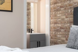 Une chambre avec un mur en briques, un lit blanc, une table de chevet grise et un miroir au-dessus du lavabo.