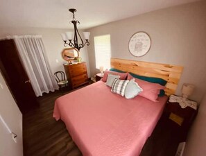 A bedroom with a wooden headboard, a pink bedspread, and decorative pillows. There is a dresser with a mirror, a chandelier, and a wall clock.