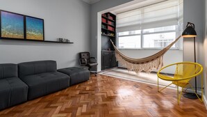 A modern living room with a herringbone wood floor, a black sofa, a yellow chair, a hammock, a bookshelf, and a large window. - Side View of the sea 2 Minutes From the Beach Fms601 (Rio de Janeiro)