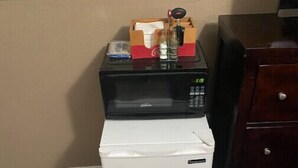 Un petit réfrigérateur, surmonté d’un four à micro-ondes, est placé à côté d’une commode en bois dans une pièce dont le sol est recouvert de planches de bois et dont le mur est de couleur beige.