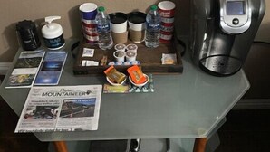 Une petite table avec un journal, des bouteilles d’eau, des tasses à café, un thermos et une machine à café portable.