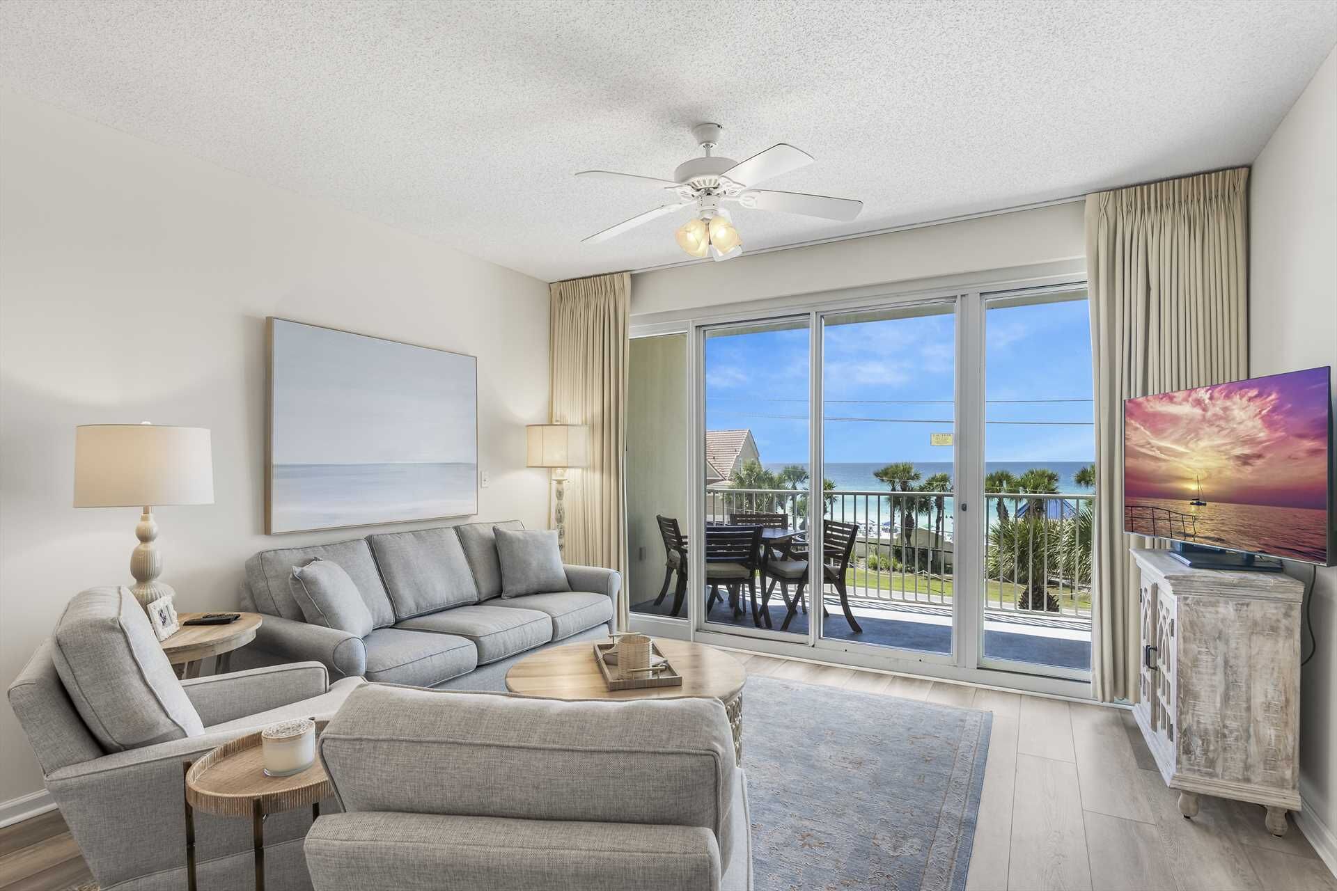 A modern living room with a grey sofa, a coffee table, and a TV. It features a ceiling fan, large windows with a view of the ocean, and a balcony with outdoor furniture.