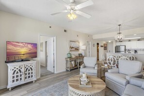 A living room with a TV, a ceiling fan, a dining area with a table and chairs, and a kitchen in the background. - New Rental | Great View | Sleeps 4| Beach Service Included (Miramar Beach FL)