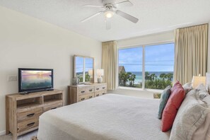Quarto de hotel com uma cama, uma televisão em uma cômoda de madeira, um ventilador de teto e uma janela grande com vista para o oceano.