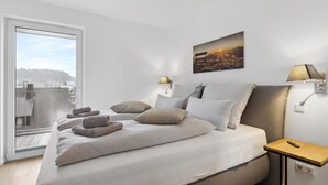 A modern bedroom with a large bed, a city view through a window, a painting of a sunset, and a bedside table with a lamp. - GebirgsResort Winterberg (Winterberg)