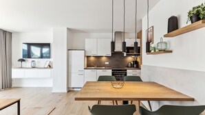Une cuisine moderne avec des meubles blancs, un coin repas avec une table en bois et des chaises vertes, et un salon avec une grande fenêtre.