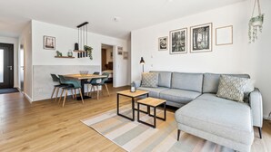 Un salon moderne avec un canapé gris, un coin repas avec une table et des chaises en bois, et une cuisine visible au fond.