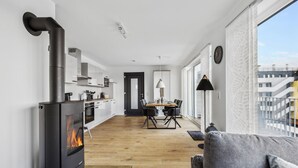 Une cuisine moderne équipée d’un poêle à bois, un coin repas avec une table et des chaises, et une vue sur un bâtiment à travers de grandes fenêtres.
