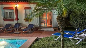 Un espace aménagé au bord de la piscine, avec des chaises longues, une petite piscine et un bâtiment dont les fenêtres sont protégées par des barreaux et qui est surmonté d’un auvent rouge.
