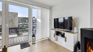 A modern living room with a large TV, a fireplace, and a balcony with a view of buildings. - GebirgsResort Winterberg (Winterberg)