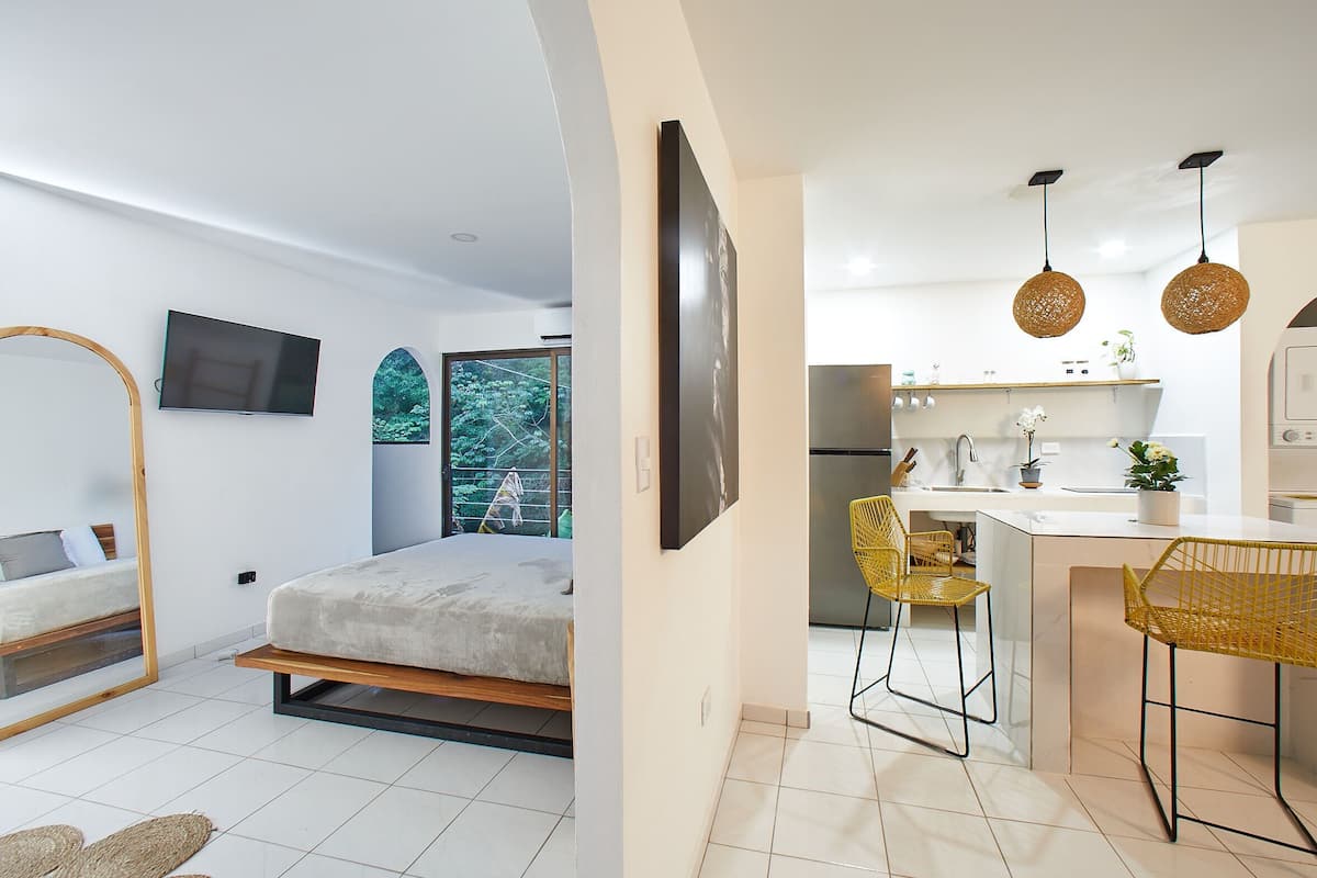 A modern bedroom with a bed, a TV, and a large window. The image also shows a kitchen with a sink, a refrigerator, and two yellow chairs.