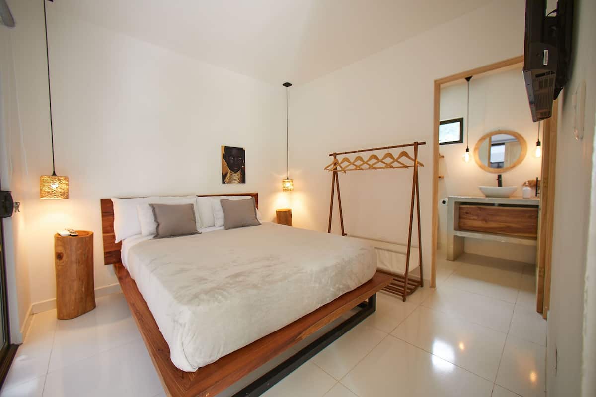 A bedroom with a wooden bed, bedside lamps, a wooden nightstand, a hanging laundry rack, and a bathroom with a sink and mirror.