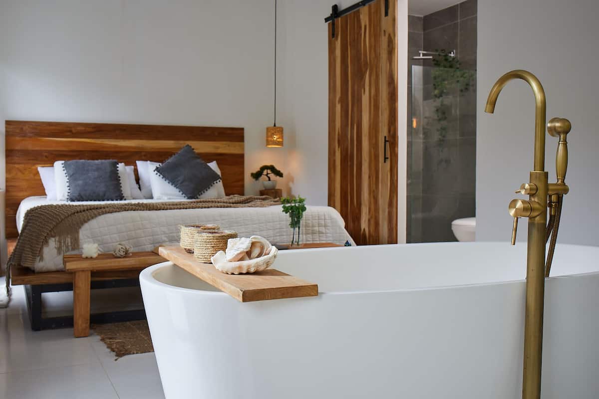 A modern bedroom with a large bed, wooden headboard, bedside lamps, and a wooden tray with bath items.