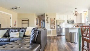 Una sala moderna con un sofá seccional de cuero, un comedor con una silla de madera, y una cocina con armarios blancos y electrodomésticos de acero inoxidable.