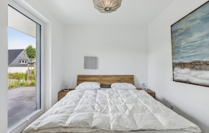 A bedroom with a large bed, a wooden headboard, a painting on the wall, and a view of a house through a window.
