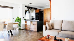 Ein modernes Wohnzimmer mit einer Couch und einem Couchtisch, ein Essbereich mit Stühlen und einer Pflanze sowie eine Küche mit Holdelementen und Edelstahlgeräten.