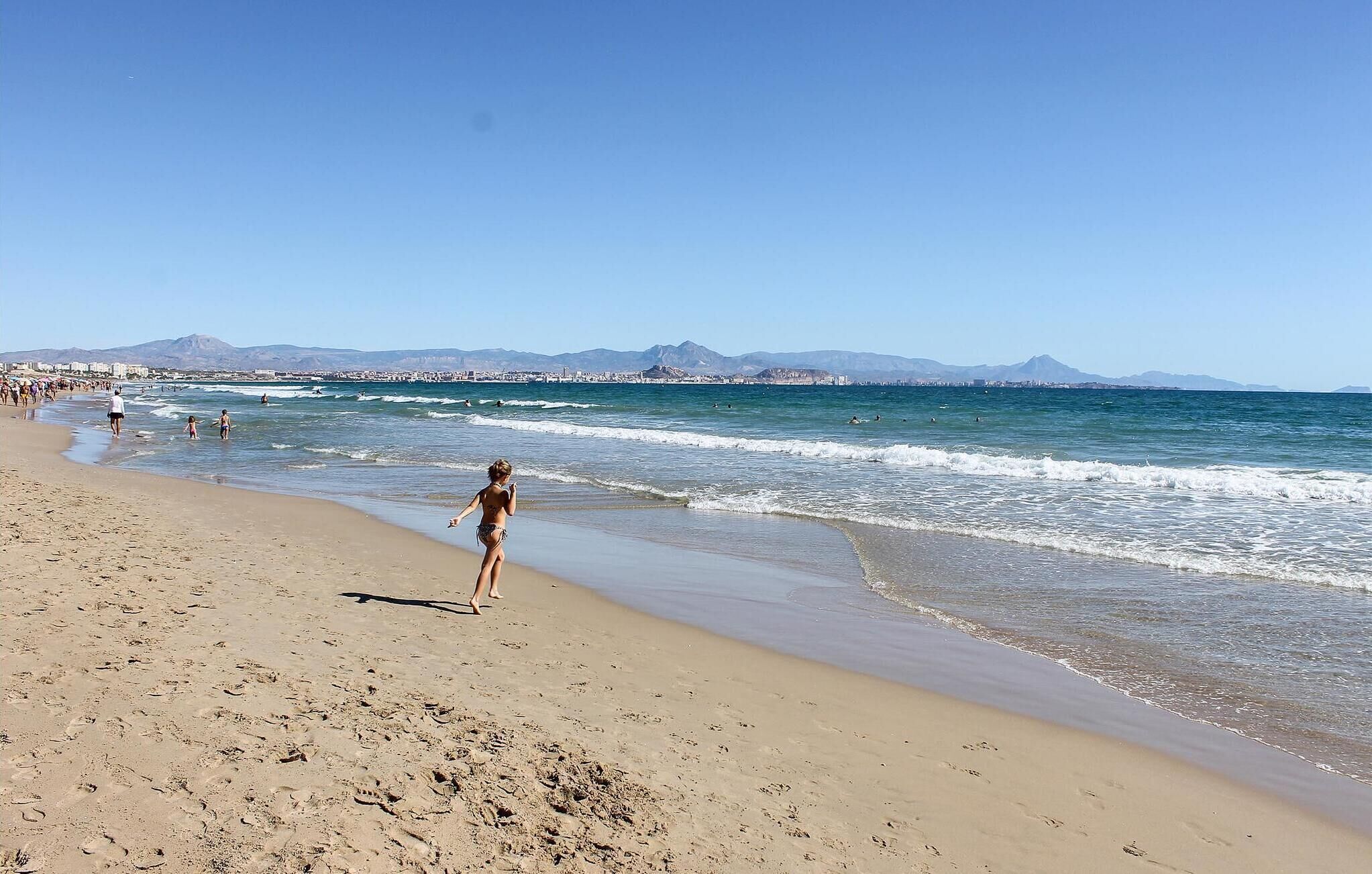 Une plage où des gens se promènent le long du rivage, des vagues qui se brisent sur le sable et, au loin, une vue sur la ville avec des montagnes en arrière-plan.