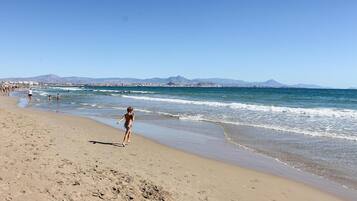 Une plage où des gens se promènent le long du rivage, des vagues qui se brisent sur le sable et, au loin, une vue sur la ville avec des montagnes en arrière-plan.