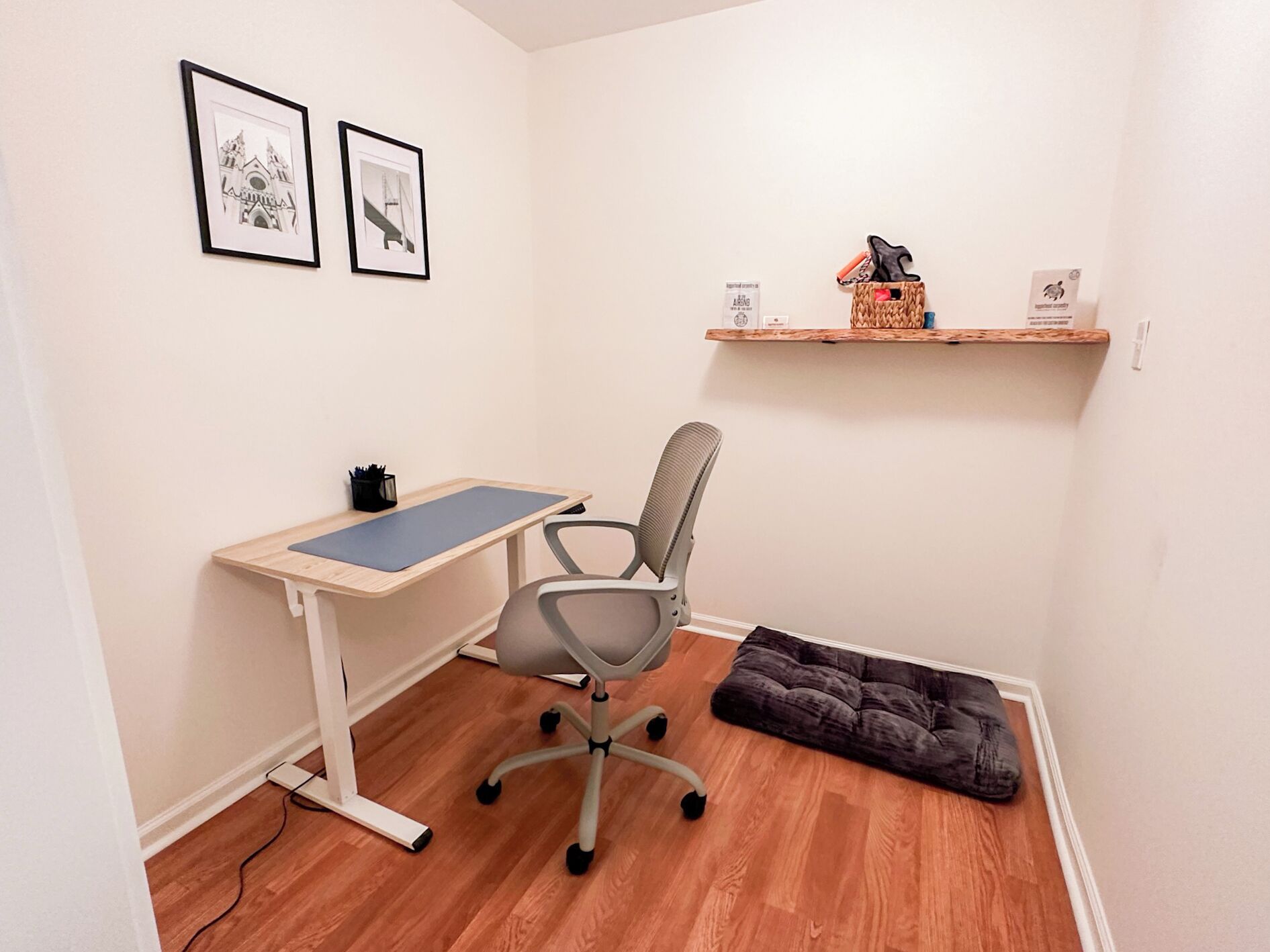 Een kleine kantoorruimte met een bureau, stoel en een met kussens beklede bank. Twee ingelijste foto's hangen aan de muur en een plank met spullen erop.